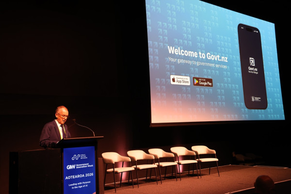 Sir Brian Roche speaking at the Public Sector Network Innovate Showcase. A presentation slide next to him displays the Govt.nz app.